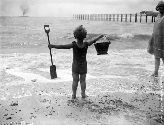 A little boy waves his bucket and spade at the waves as he watches the sea on the beach, 1927
