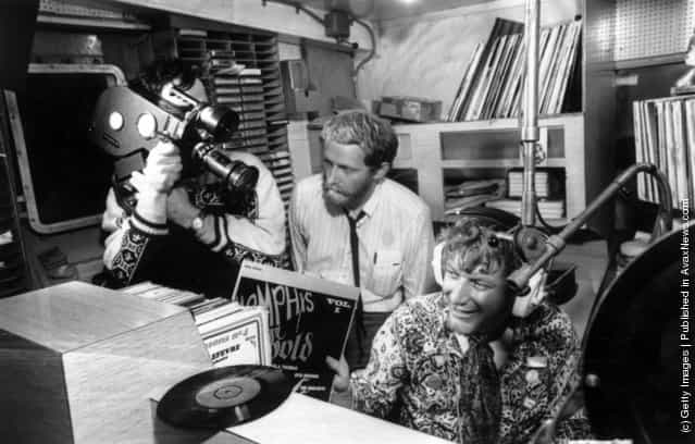 The World in Action team making a program about the pirate radio ship Caroline, filmed by Paddy Searle, and produced by Mike Hodges. The DJ being filmed is Robbie Dale, and Hodges is standing behind him