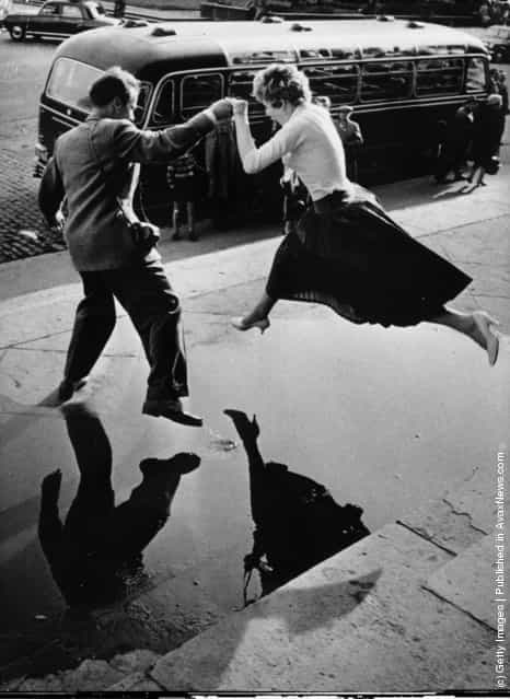 A man gives a woman a helping hand as she takes a flying leap over a large puddle on the pavement
