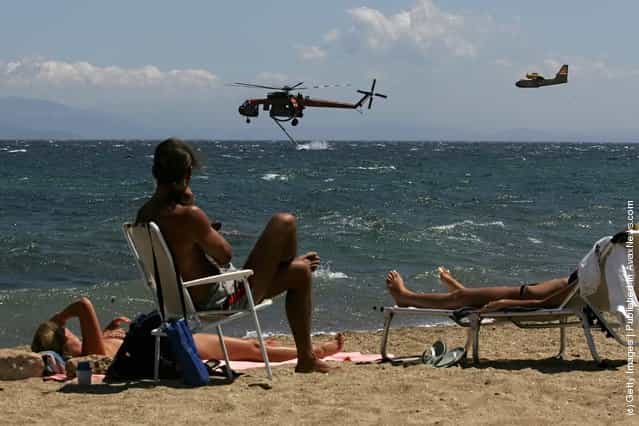 Sunbathing tourists watch a helicopter as it collects sea water to dowse fire, as wild fires burn in the suburbs