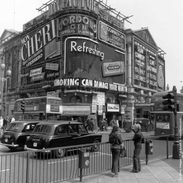 1971: Two girls pointing at the signs in Piccadilly circus, London, which include an advertisement for Wrigleys Chewing gum and a Government health warning about smoking