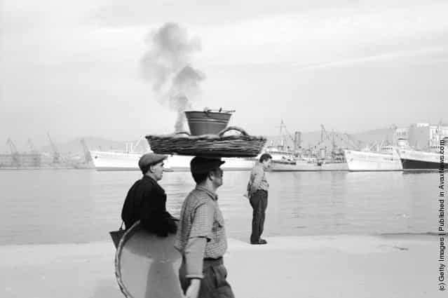 1947: A man balancing a large basket and a pail on his head walks along the harbour at Piraeus in Greece