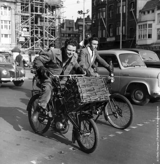1955: Two young men cycling amongst traffic in Holland, one on a bicycle with a motor attached and a large basket at the front