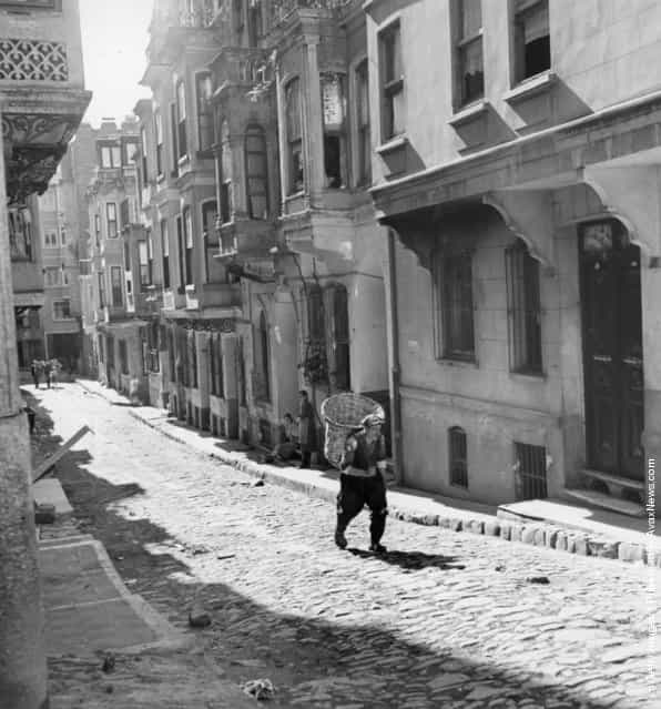 1957: An old man carrying a large basket walks down a narrow cobbled street in what is by night Istanbuls main area for prostitution
