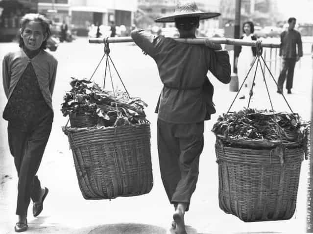 1960: In a Kowloon street a bare-footed woman carries two large panniers of vegetables on a yoke across her shoulders