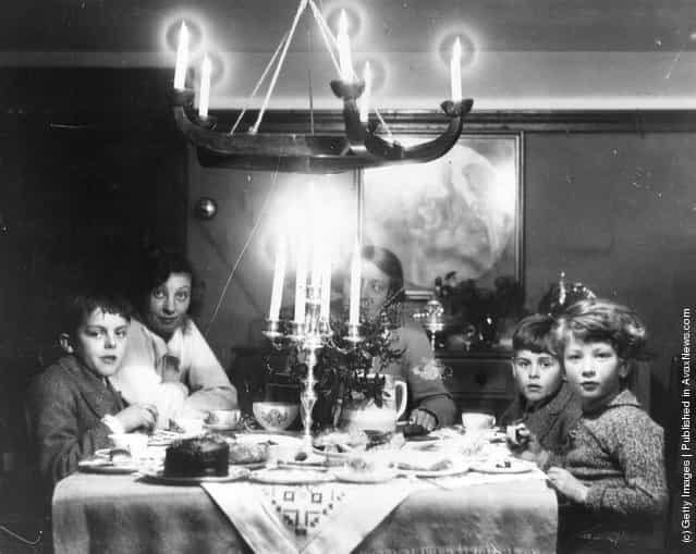 1935: A family at Christmas tea time with chocolate cake, a candelabra and holly on the table