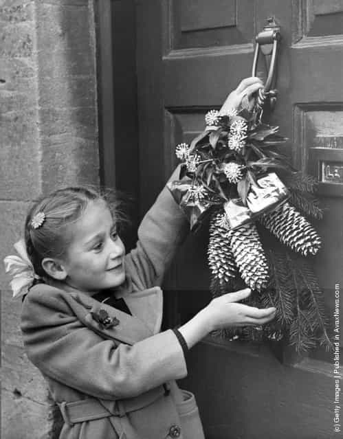 1954: A young girl hangs a Christmas garland on the door of her cottage home at Northleach, Gloucestershire, in accordance with an old local custom