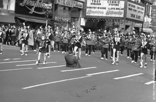 A man squats in the street to photograph a brass band and majorettes marching in the Thanksgiving Parade near Times Square, New York, 1961