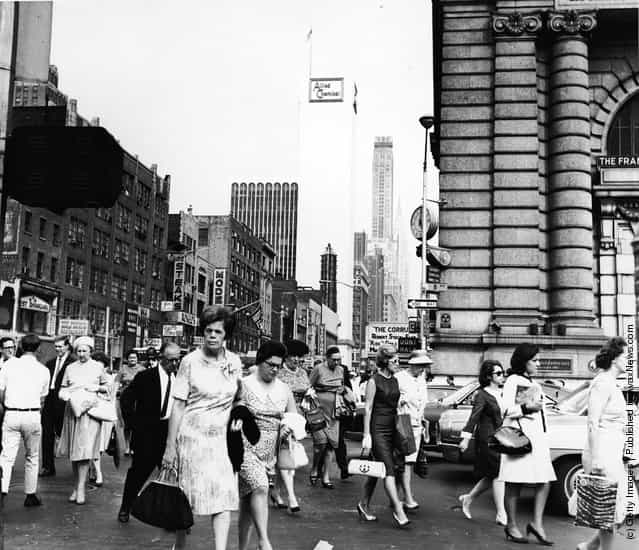 Crowds of pedestrians walk at the intersection of Eighth Avenue and 42nd Street, with the Allied Chemical building visible in the background, New York City, circa 1962
