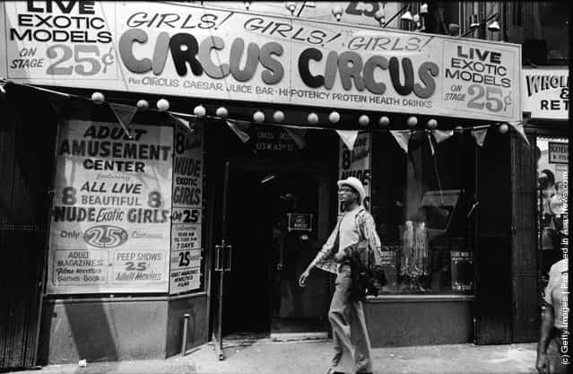 Exterior view of Circus Circus, an adult amusement center, in Times Square as a man walks past wearing a hat and carrying his coat over his arm, New York, New York, 1970s. The marquee advertises exotic models, nude girls, books, magazines, games, and peep shows, as well as a juice bar and high-potency protein health drinks