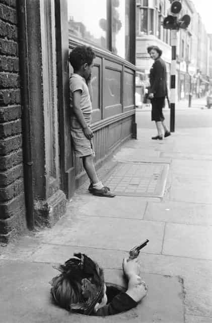 A young boy wearing an Indian headdress hides in a coal hole and takes aim with a toy pistol, London, 7th August 1954. (Photo by Thurston Hopkins/Picture Post)