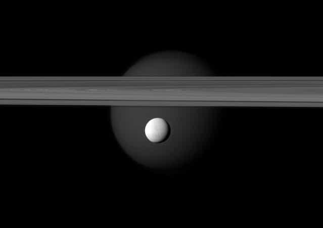 A brightly reflective Enceladus appears before the rings of Saturn, while the planet's larger moon Titan looms in the distance. Enceladus (313 miles, or 504 kilometers across) is in the center of the image. Titan (3,200 miles, or 5,150 kilometers across) glows faintly in the background beyond the rings, in this image acquired by NASA's Cassini spacecraft on March 12, 2012. (Photo by NASA/JPL-Caltech/Space Science Institute)