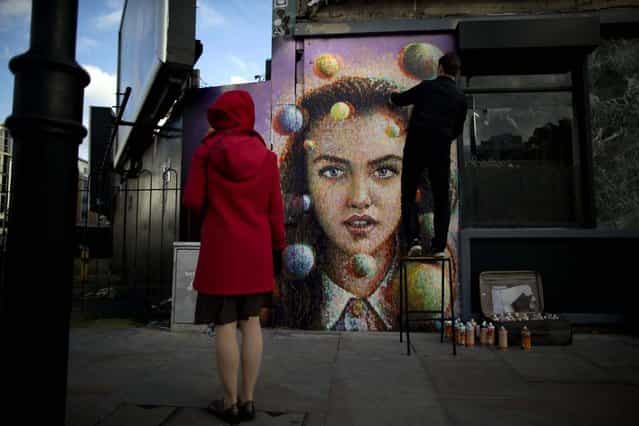 A passerby watches Australian artist James Cochran, also known as Jimmy C, put finishing touches to his spray painted 3D piece [Girl with Spheres in 3D] on the outside wall of a cafe in east London, November 22, 2012. The effects of the 3D painting can only be seen when the viewer looks through a pair of 3D glasses, which the cafe will keep a stock of to lend to people. (Photo by Matt Dunham/Associated Press)