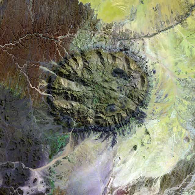 Over 120 million years ago, a single mass of granite punched through the Earth’s crust and intruded into the heart of the Namib Desert in what is now northern Namibia. Today the mountain of rock called the Brandberg Massif towers over the arid desert below. A ring of dark, steep-sided rocks forced upward during the mountain’s arrival encircles the granite intruder. Its volcanism has long since stilled, but the granite core left behind apparently glows red in the light of the setting sun. The formation is a remnant of a long period of tumultuous volcanic and geologic activity on Earth during which the southern super-continent of Gondwana was splitting apart. The mountain influences the local climate, drawing more rain to its flanks than the desert below receives. The rain filters into the mountain’s deep crevices and slowly seeps out through springs. Unique plant and animal communities thrive in its high-altitude environment, and prehistoric cave paintings decorate walls hidden in the steep cliffs that gouge the mountain. To the southwest of Brandberg Massif, an older and more-eroded granite intrusion blends in subtly with the desert landscape, while along the Ugab River at upper left, cracks line the brown face of an ancient plain of rock transformed into gneiss by heat, pressure, and time. This image was acquired by Landsat 7’s Enhanced Thematic Mapper plus (ETM+) sensor. (Photo by NASA/GSFC/USGS EROS Data Center)