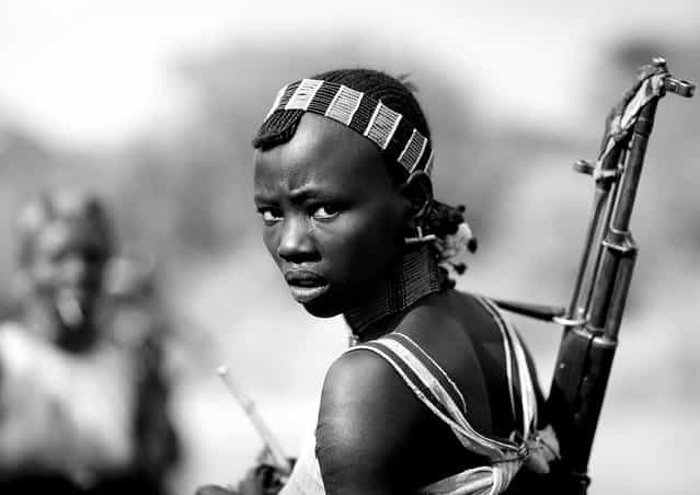 [Hamer girl with gun – Omo Ethiopia. During bull jumping ceremony, Hamer girls act like men, and even more, to show that they do not fear anything. So they drink a lot, and they dance with guns. Very impressive ceremony!
The Hamar is a catlle herder tribe which lives on the Eastern side of the Omo Valley in Southern Ethiopia. Honey collection is their major activity and their cattle is the meaning of their life. There are at least 27 words for the subtle variations of colours and textures of a cattle ! And each man has three names: a human, a goat and a cow name.
The Hamar have very unique rituals such as a bull-leaping ceremony, that a young men has to succeed in order to get married. The cow jumping is an initiation rite of passage for boys coming of age in Hamar tribe. Cows are lined up in a row. The initiate, naked, has to leap on the back of the first cow, then from one bull to another, until he finally reaches the end of the row. He must not fall of the row and must repeat successfully the test four times to have the right to become a husband. While the boys walk on cows, Hamar women accompany him: they jump and sing. Totally committed to their initiated sons, the mothers are whipped to blood, in order to prove their courage and accompany their sons during the test]. (Photo and comment by Eric Lafforgue)