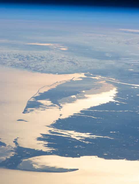 The coastline of the northeastern U.S., observed from the ISS on February 14, 2013. The Atlantic Ocean, including Cape Cod Bay and Buzzards Bay along the coastlines of the states of Massachusetts and Rhode Island has a burnished, mirror-like appearance in this image. This is due to sunlight reflected off the water surface back towards the astronaut-photographer. The peak reflection point is towards the right side of the image, lending the waters of Long Island Sound (at image center, to the north of Long Island) and the upper Massachusetts coastline an even brighter appearance. Sunglint also illuminates surface waters of Chesapeake Bay (top center) located over 400 km to the southwest of the tip of Long Island. The high viewing angle from the ISS also allows the Earth's curvature, or limb, to be seen, and blue atmospheric layers gradually fade into the darkness of space across the top part of the image. (Photo by NASA/The Atlantic)