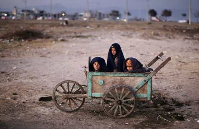 Afghan refugee boys wrap themselves with blankets to avoid the evening cold while sitting in a wooden cart as they look at a group of girls playing hopscotch, not pictured, in a field on the outskirts of Islamabad, on November 30, 2012. (Photo by Muhammed Muheisen/AP Photo)