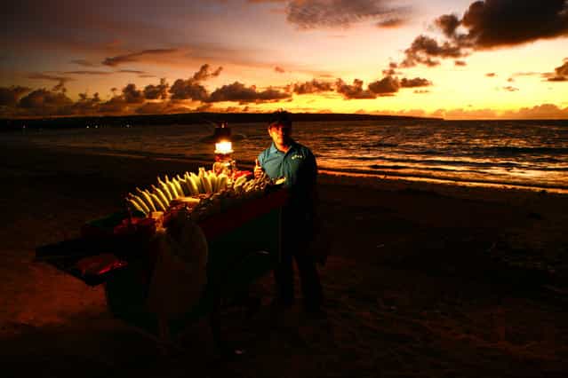 [Indonesian Man Selling Corn]. This man waited patiently on Jimbaran Beach in Bali, Indonesia to sell corn to tourists after they ate their candlelit dinners on the beach. I waited and watched how the gorgeous sunset slowly started to match the lighting of his cart of corn and then walked over and asked him to take this portrait of him. I love how he gave me the [thumbs up]. (Photo and caption by Laura Grier/National Geographic Traveler Photo Contest)