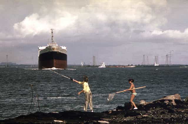 A freighter moves slowly up the Houston ship channel as girls fish from the shore, September 1973. Oil derricks are visible in the background. (Photo by Blair Pittman/NARA via The Atlantic)