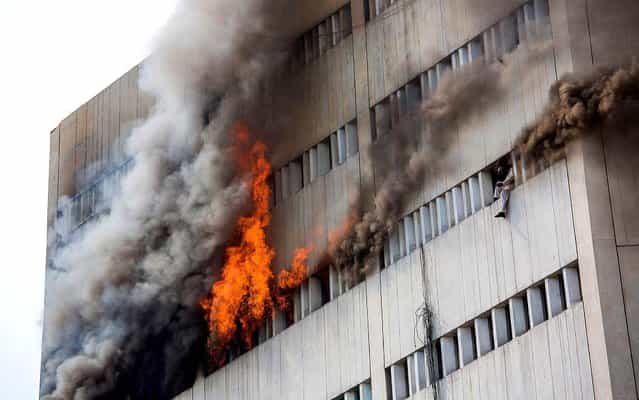 As a fire burns at the Lahore Development Authority Plaza, a man is perched at a window moments before being overcome by fumes and falling out of the window. Rescue work is underway at the site of the fire as helicopters are used to save stranded victims from the roof of the building. (Photo by Daniel Berehulak/Getty Images)
