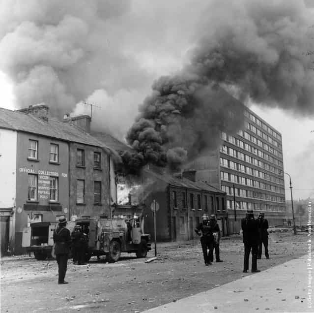 1969: Heavy black smoke billows from a building after a bomb attack in the Bogside area of Belfast. Firemen are in attendance