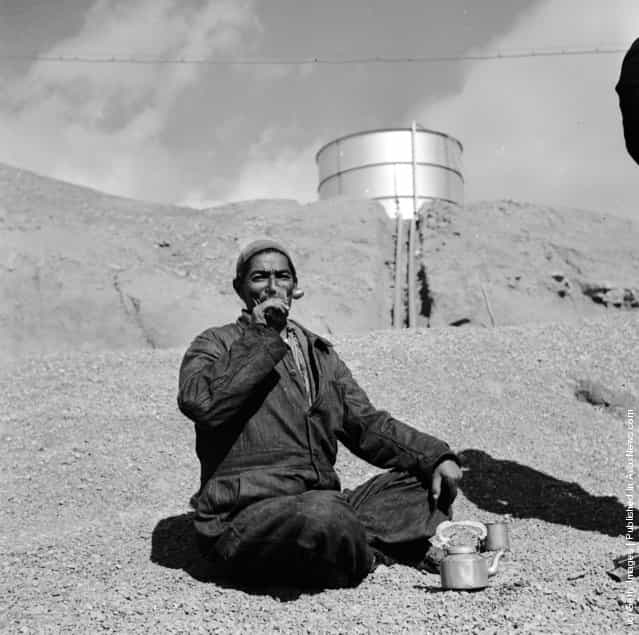 1954: An Iranian oil worker in an arid landscape, an oil tank in the distance, sits enjoying the labourers staple diet of bread and tea