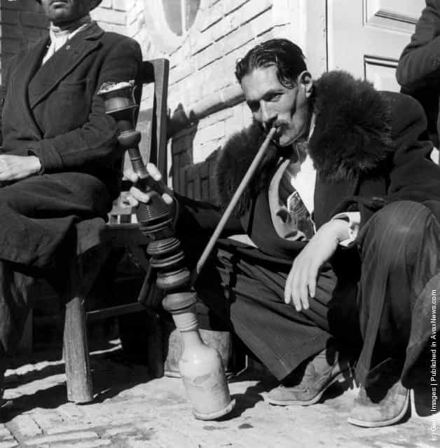 1955: An Iranian smokes a Persian hookah or water pipe on the pavement in Isafahan. The pipe is hired by a specialist merchant for single use