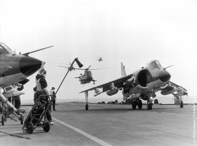 A Sea Harrier jump jet on the flight deck of HMS Hermes heading to the Falklands after the Argentinian invasion of the islands, April 1982. A Sea King helicopter hovers in the background
