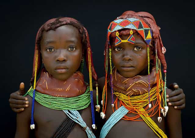 [Two Mumuhuilas little girls – Angola. Those little girls from the Mumuhuila tribe live near Chibia, in the south of Angola. They wear the traditional hairstyle and the big necklace. The necklace shows if they are teens or not. They still are children as the necklace is red. On their hair, they wear the traditional hairstyle, made with a mix of trunk, oil and cow dung. It was impossible to make them smile as pausing for a white man was too impressive for them, it was the first time they met tourist. Imagine the face they had when i gave them a polaroid at the end of the session! Those little girls live dressed like that, they were not dressed for me]. (Eric Lafforgue)