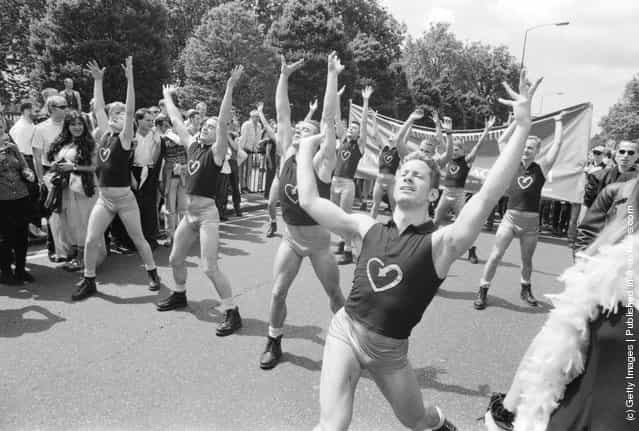 Male dancers standing with their arms raised above their heads, wearing black t-shirts with a heart logo, during the Gay Pride parade in London, England