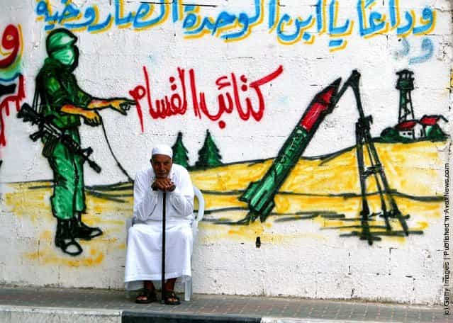 An elderly Palestinian man sits in front of a wall with a mural showing a masked militant from Ezz El-Deen Al-Qassam Martyrs Brigade, the military wing of the Islamic resistance movement Hamas, as he fires a homemade Qassam rocket towards Israeli settlements, in Jabaliya refugee camp northern Gaza Strip August 28, 2003