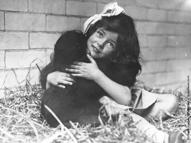 circa 1930: A little girl with a ribbon in her hair cuddles a monkey