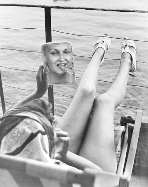 1949: Feet up on the rails, a woman studies her reflection in a mirror whilst sunbathing on the deck of a yacht in Portofino, Italy