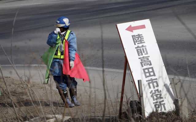 In this Friday, February 22, 2013 photo, standing by a signboard reading: [Rikuzentakata City Hall] a worker holds flags to control traffic under cold weather in a street near a new road construction site in Rikuzentakata, Iwate Prefecture. Japan's progress in rebuilding from the tsunami that thundered over coastal sea walls, sweeping entire communities away, is mainly measured in barren foundations and empty spaces. Clearing of forests on higher ground due to be leveled to make space for relocating survivors has barely begun. Japan next week, will observe two years from the March 11, 2011 disasters which devastated in the northeastern Pacific coast of the country. (Photo by Junji Kurokawa/AP Photo)