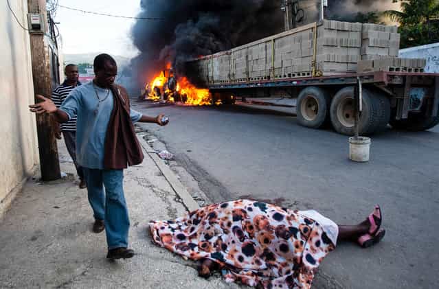 A resident expressing shock, walks by the body of rice vendor Dasilia Daniel, who was accidentally shot dead during a road rage incident as she walked to work in the Petionville suburb of Port-au-Prince, Haiti, Friday, July 19, 2013. According to a cousin of Daniel, the driver of a semi-trailer truck tapped the back of a pickup, enraging its driver, who then stepped out from his vehicle, gun in hand, and shot at the semi-truck driver, missing him and killing Daniel. Fellow vendors set the vehicles on fire in retaliation for the death of their friend. (Photo by Dieu Nalio Chery/AP Photo)