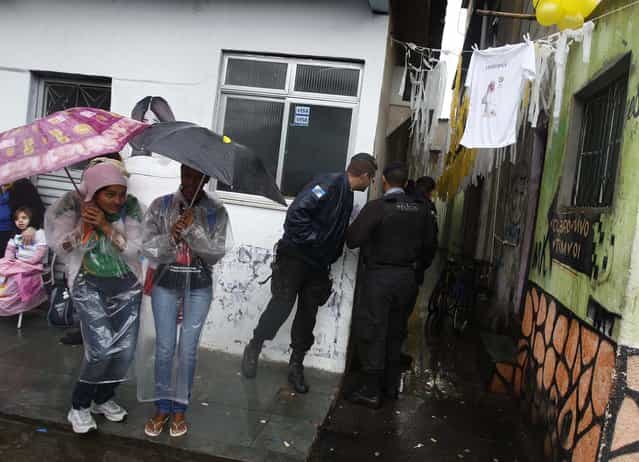 Residents of the Varginha slum wait under umbrellas, while police guard the street, as they wait for Pope Francis to visit the slum on July 25, 2013. (Photo by Pilar Olivares/Associated Press)
