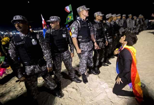 A protestor wearing a gay pride flag kneels in front of a line of National Guard officers preventing demonstrators from approaching the site of a vigil with Pope Francis during World Youth Day celebrations on Copacabana beach. Demonstrators were protesting for women's rights and gay rights, as well as against government corruption and the World Cup. (Photo by Silvia Izquierdo/Associated Press)