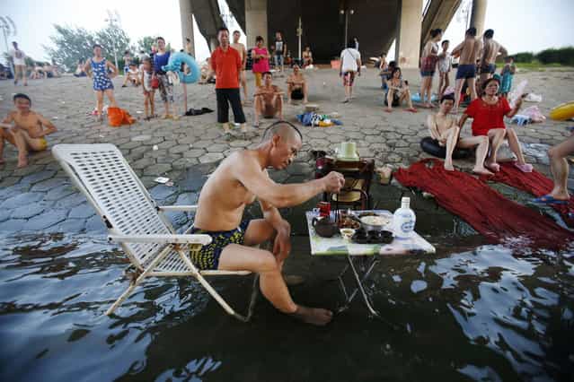 A man sitting on a beach chair in his swimming trunks takes his meal at a small table partially dipped into the Hanjiang river to escape the summer heat, as other swimmers look on under a bridge in Wuhan, Hubei province July 28, 2013. The highest temperature in Wuhan reached 37 degrees Celsius (98.6 degrees Fahrenheit) on Sunday, local media reported. Picture taken July 28, 2013. (Photo by Reuters/China Daily)