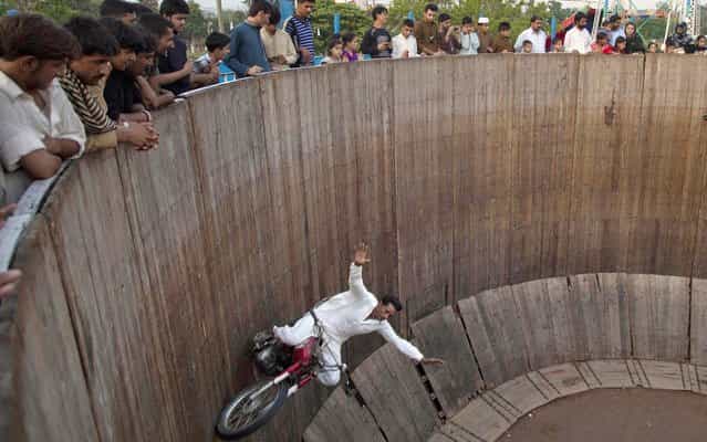 People watch a man demonstrate his motorcycle skills inside the [well of death] at a fair set up on the occasion of the Eid al-Fitr holiday in Rawalpindi, Pakistan, Monday, August 12, 2013. (Photo by B. K. Bangash/AP Photo)
