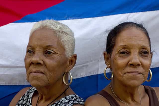 In this September 29, 2013 photo, 65-year-old twin Fe Fernandez, left, and her sister Esperanza pose for a portrait in front a Cuban flag on their street in Havana, Cuba. Neighbors all marvel at the 12 sets of twins living along two consecutive blocks in western Havana, ranging in age from newborns to senior citizens. [We were the first ones], said Fe Fernandez. (Photo by Ramon Espinosa/AP Photo)