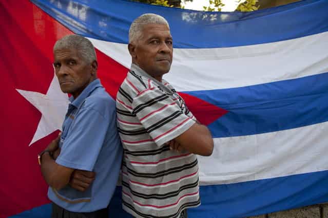 In this September 29, 2013 photo, twins Orlando Gonzalez, left, and Roberto Gonzalez pose for a portrait in front a Cuban flag along their street in Havana, Cuba. The Gonzalez brothers are one of 12 sets of twins living along two consecutive blocks in western Havana, ranging in age from newborns to senior citizens. (Photo by Ramon Espinosa/AP Photo)