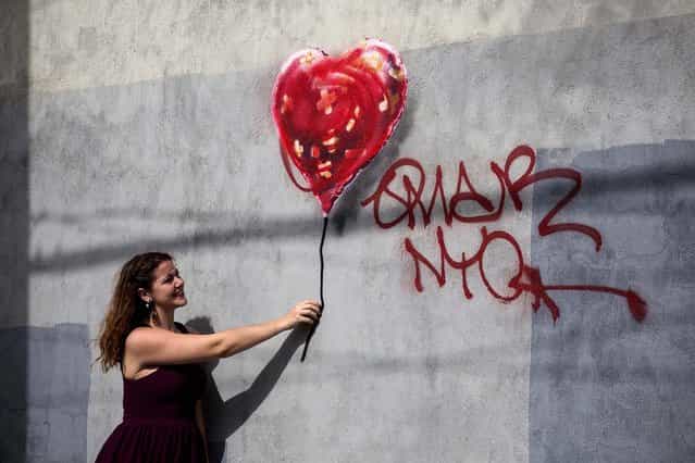 A woman poses with a Banksy piece depicting a heart-shaped balloon covered in bandages, on October 7 in the Red Hook neighborhood of the Brooklyn borough of New York City. The piece was defaced with red spray paint shortly after being completed. (Photo by Andrew Burton/Getty Images)