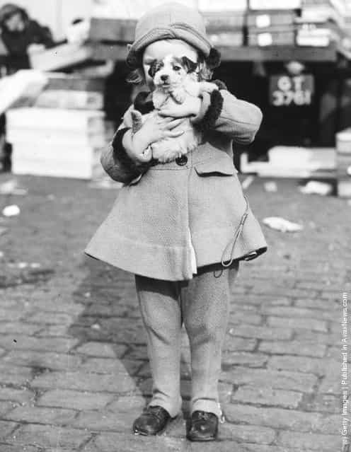 A little girl carries a struggling puppy away from a market where she has just bought him, 1938